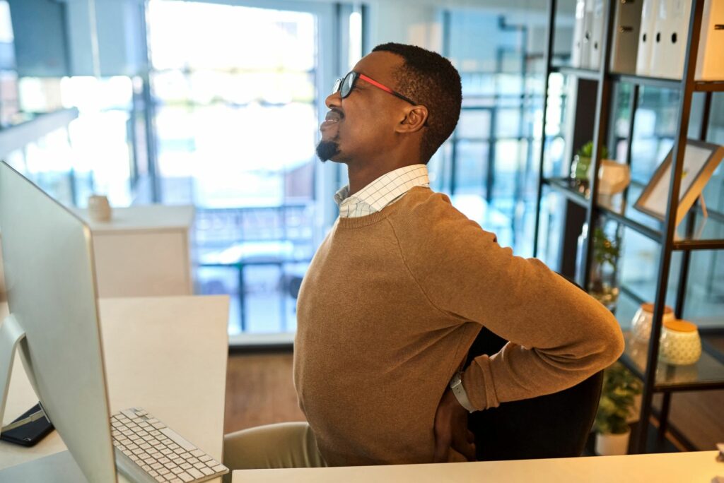 Here a man winces and reaches for his back while sitting at his work cubicle. Office spaces are designed to fit our bodies. What about driving ergonomics?