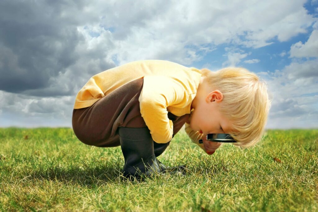A curious little boy bends over to look at the blades of grass through a magnifying glass.