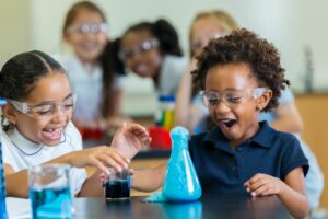 Two kids working on a classroom chemistry experiment are overjoyed as blue foam rises to the top of a beaker. Lab partners work together.
