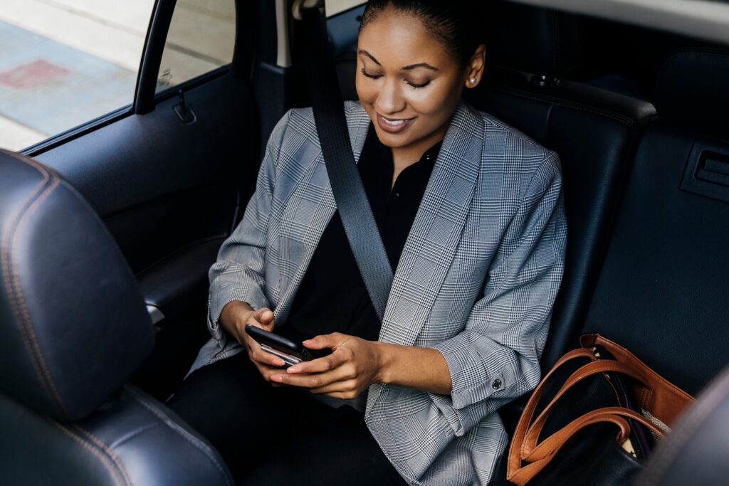 A woman is pictured here safely buckled up in the back seat of a rideshare vehicle. She is scrolling through information on her phone.